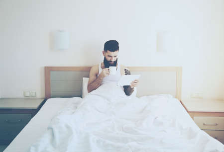 Bearded man lying in morning bed with coffee or tea cup and digital tablet using app or reading news feedの写真素材