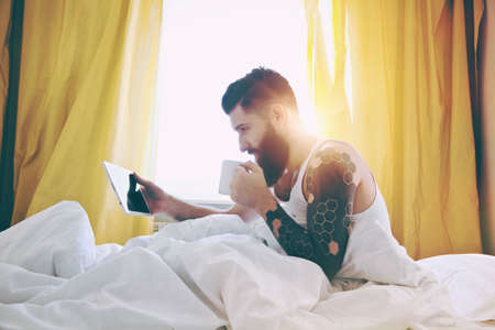 Bearded man lying in bed with morning coffee and digital tabletの写真素材