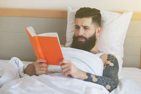 Bearded man lying in morning bed and reading bookの写真素材