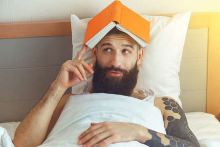 Bearded man lying in morning bed with bookの写真素材