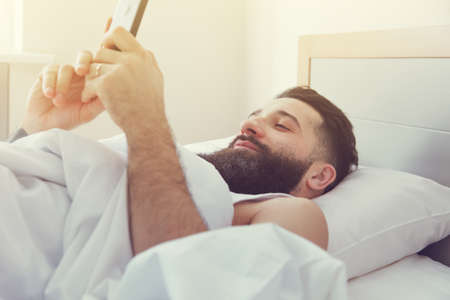 Bearded man lying in morning bed with phone using app or reading news feedの写真素材