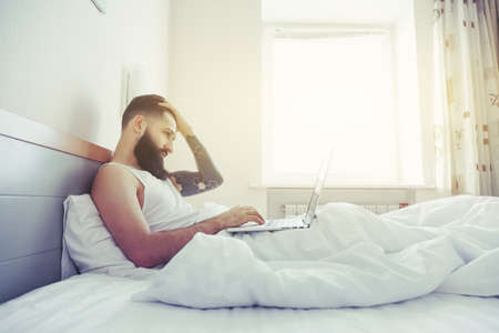 Bearded man lying in morning bed working with laptopの写真素材