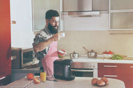 bearded man drinking cup of morning coffee with coffee machine while breakfast at kitchenの写真素材