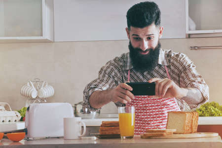 funny bearded man making photo with camera phone of morning breakfast at kitchenの写真素材