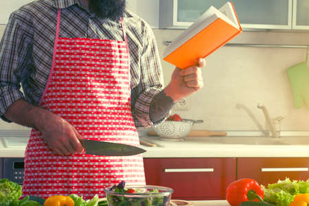 man cooking at kitchen making healthy vegetable salad and reading recipe bookの写真素材
