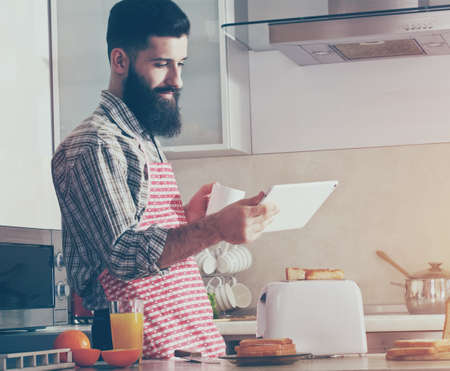 bearded man drinking morning coffee or tea and using digital tablet at kitchenの写真素材