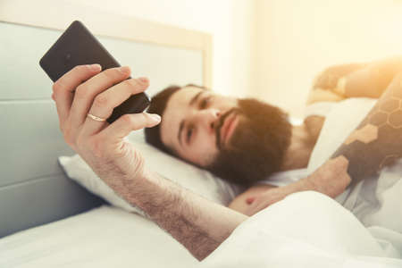 Bearded man lying in morning bed with phone using app or reading news feedの写真素材