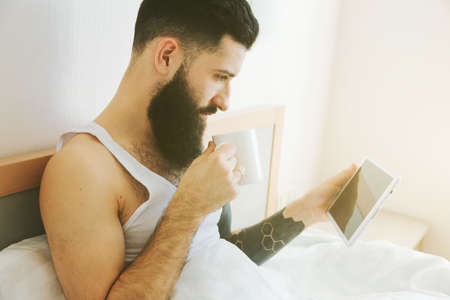 Bearded man lying in morning bed with coffee or tea cup and digital tablet using app or reading news feedの写真素材