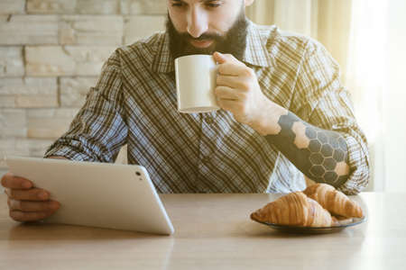 Smiling bearded man drinking cup of morning coffee or tea with digital tabletの写真素材