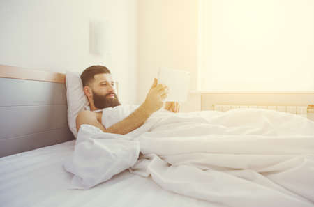 Bearded man lying in morning bed with digital tablet using app or reading news feedの写真素材