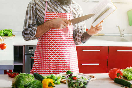 man cooking at kitchen making healthy vegetable salad and reading recipe bookの写真素材
