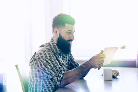 Smiling bearded man drinking cup of morning coffee or tea with digital tabletの写真素材