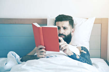 Bearded man lying in morning bed and reading bookの写真素材