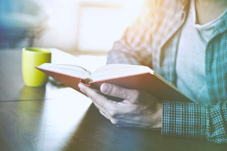 man reading book with cup of tea or coffee at home in morning sunlight