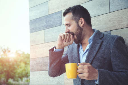 businessman yawning and holding morning coffee at wall backgroundの写真素材