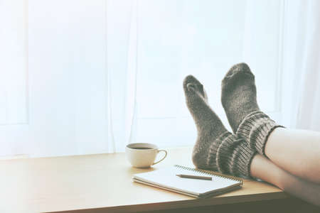 Woman resting keeping legs in warm socks on table with morning coffee and notebook. Space for textの写真素材