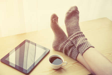 Woman resting keeping legs in warm socks on table with morning coffee and reading e-book or tabletの写真素材