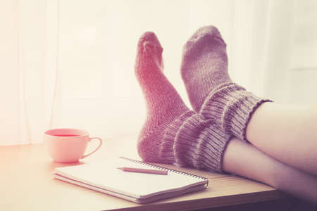 Woman resting keeping legs in warm socks on table with morning coffee and notebookの写真素材