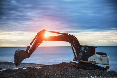 backhoe or digger working with bucket at industrial earth excavation site near sea in sunrise lightの写真素材