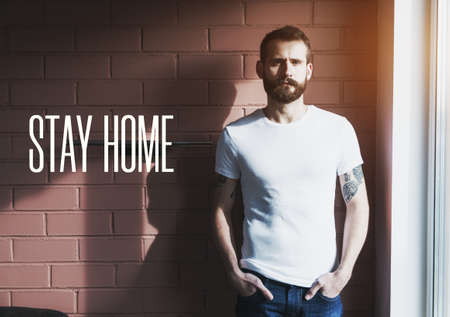 Handsome bearded man in white t-shirt on brick wall background and text Stay home. Home isolation and quarantine during coronavirus covid-19 pandemic.の写真素材