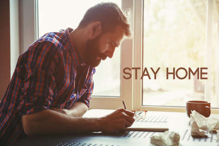 Man writing in notebook with pen and text Stay home. Home isolation and quarantine during...