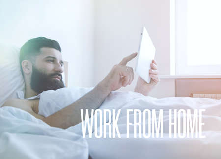 Bearded man lying in morning bed with digital tablet using app or reading news feedの写真素材