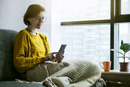 Woman working with phone at home quarantine. Coffee or tea and warm plaid for comfortable workplace. Stay at home campaign.の写真素材