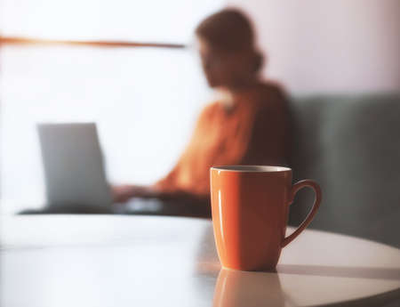 Coffee or tea mug with woman working with laptop at home background. Comfortable workplace on couch. Space for textの写真素材