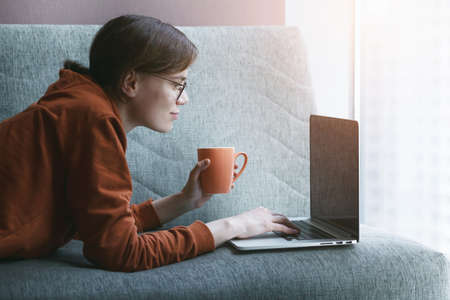 Woman working with laptop at home. Comfortable online workplace with coffee on couch.の写真素材