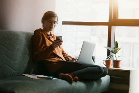 Woman working with laptop and phone at home. Comfortable workplace with coffee on couch. Stay at home campaign.の写真素材