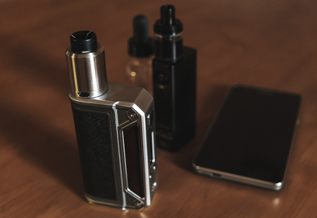 Vape devices, mods, dripping atomizer, tank, E-cigarette for vaping, liquid in the bottle and mobile phone on wooden tableの写真素材