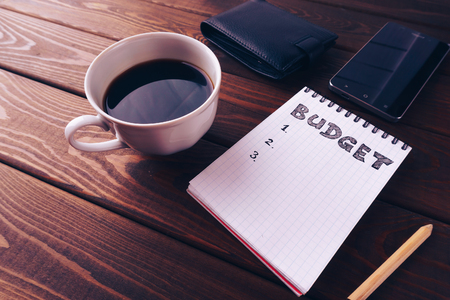 Budget list on notepad, mobile phone, pouch and cup of coffee on dark wooden table. Budget planing concept. Vintage style, retro toned picture.の写真素材