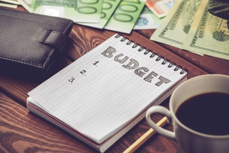 Budget planing concept. Notepad with word Budget, mobile phone, cup of coffee, pouch and money on wooden background. Write idea success solution. Vintage toned picture.の写真素材