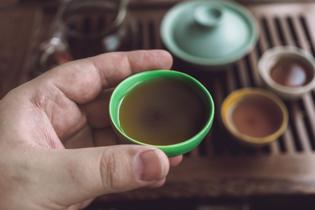 Male hand holding piala or bowl with Shu Puerh on blurred background of Asian tea set, Traditional Chinese Tea Ceremonyの写真素材