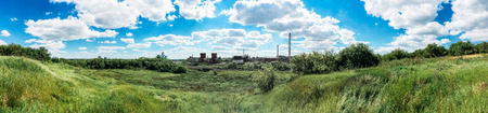 Panorama of green hills and industrial plant or factory with chimneys or pipes on a sunny dayの写真素材