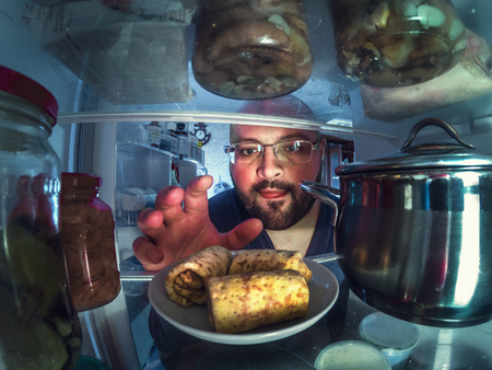 Hungry young man takes plate with pancakes from open fridge or refrigerator, toned photo