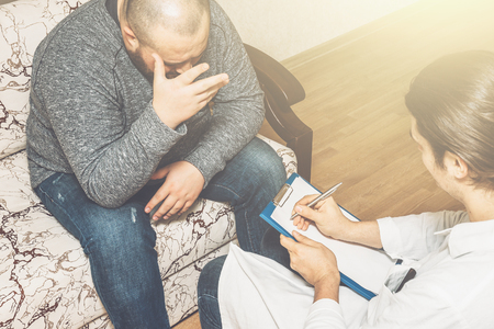 Young man at psychotherapist consulting, sits on couch, emotionally speaks about his problems, doctor makes notes. Psychologist help session concept, tonedの写真素材