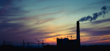 Panorama of city sunset and silhouettes of cranes, high-rise buildings and construction site with smoke, tonedの写真素材