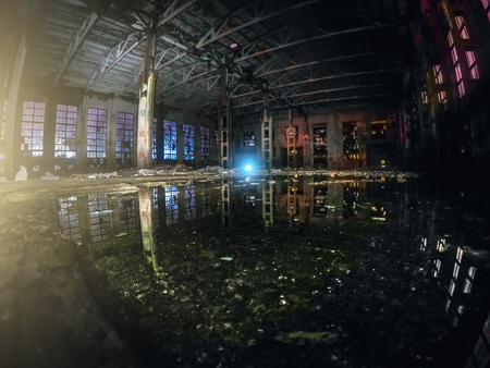 Large empty abandoned warehouse building or factory workshop at night with reflection in water and light effects, abstract ruins backgroundの写真素材