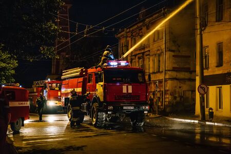 Rybinsk, Russia -31 July 2018 : Firefighters with fire hose and fire engines or trucks are fighting fire in residential building at night. Huge smoke, hot flame in burning houseのeditorial素材