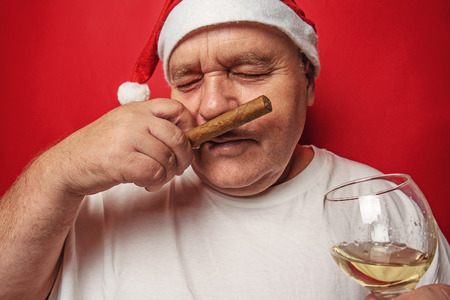 Funny drunk mature man in red Christmas hat holding glass with alcohol, sniffing cigar and enjoying, drinking in New Year holidays concept.の写真素材