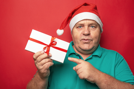 Portrait of man in red Christmas hat pointing with finger on Gift Box on red background, New Year presents for family conceptの写真素材