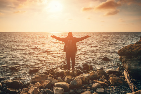 Silhouette of man with open arms or hands standing on sea beach at sunset rays. Freedom and relax in...