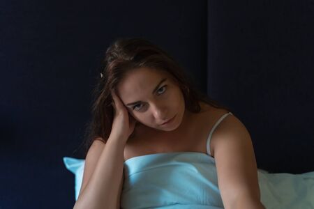 Tired young girl sitting in bed and looks at camera, insomnia, stress and sleep disorder concept.
