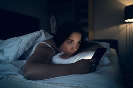Insomnia and social media addiction concept. Young woman uses smartphone while lying in bed at night, tonedの写真素材