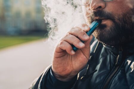 Man smokes new Vape Pod System, inhales and exhales vapor of electronic cigarette, vaping concept,...
