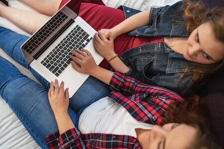 Lesbian couple or two female friends using laptop in big bed, smiling and talking to each other, top...