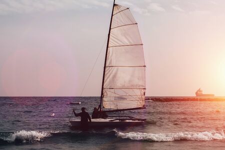 Boat with sail and silhouettes of people in sea water at sunset. Outdoor vacation in mediterranean resort.の写真素材