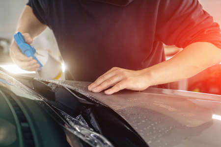Detailer worker sprays by soap sud and wrap PPF or car protection film on vehicle hood.の写真素材