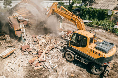 Demolition of building by industrial excavator. Bucket breaks walls and roof of old house full of dust, aerial view.の写真素材